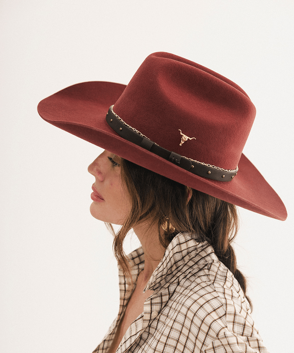 A woman in profile wearing a wide-brimmed felt cowboy hat in a deep reddish-brown color, featuring a black band with gold studs and a small gold longhorn emblem. She is wearing a beige and brown plaid shirt against a clean white background #color_rusty red