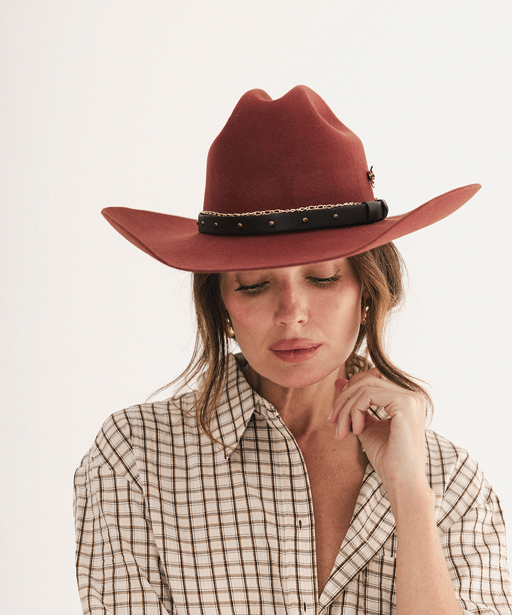 A woman wearing a wide-brimmed rust-colored felt cowboy hat with a black band and gold chain detail, paired with a beige and brown plaid button-down shirt, set against a plain white studio background #color_rusty red