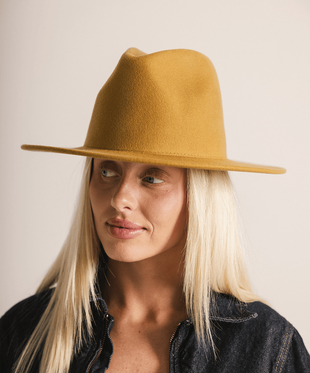 Woman wearing a mustard colored fedora hat and a black coat against a plain background #color_mustard