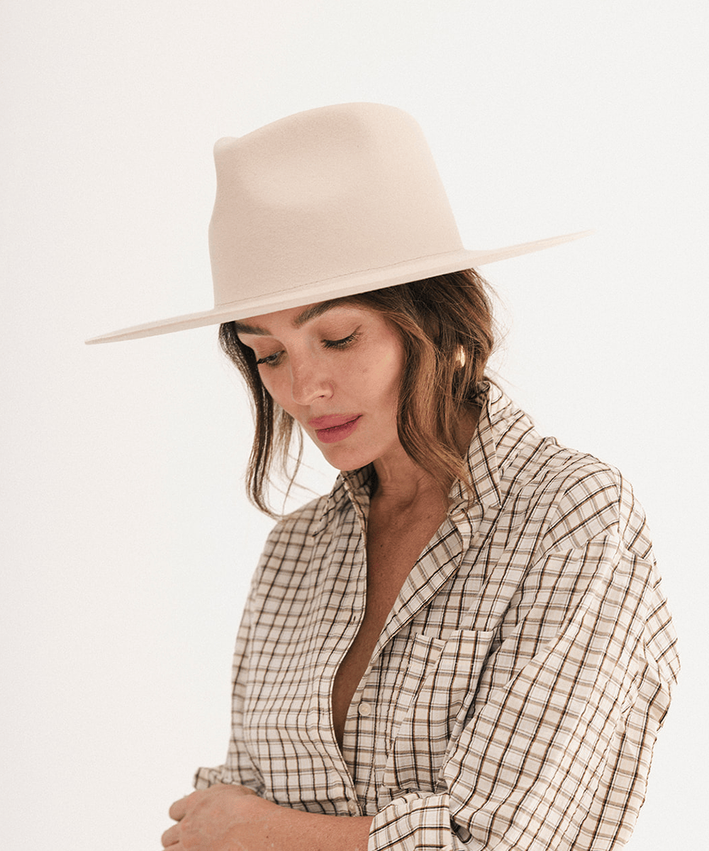 A woman wearing a wide-brimmed beige fedora hat and a beige-and-white checkered button-down shirt, photographed against a clean white studio background #color_cream