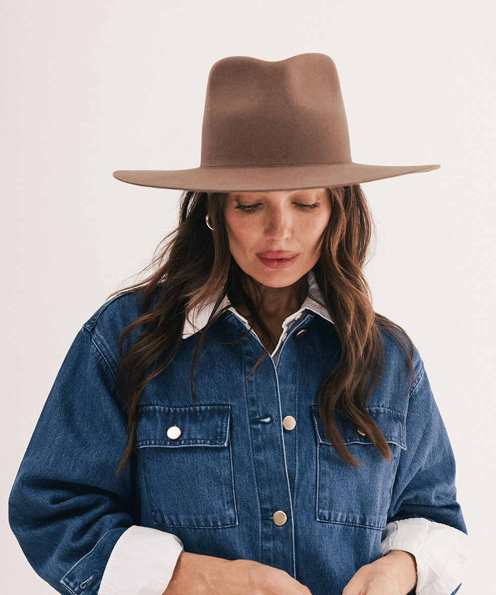 A woman wearing a brown wide-brim felt hat and a blue denim jacket over a white collared shirt, photographed against a plain light background #color_chocolate
