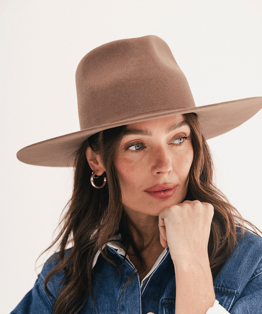 A woman wearing a wide-brimmed felt hat in a warm taupe-brown color, paired with a denim shirt and gold hoop earrings, set against a plain off-white background #color_chocolate