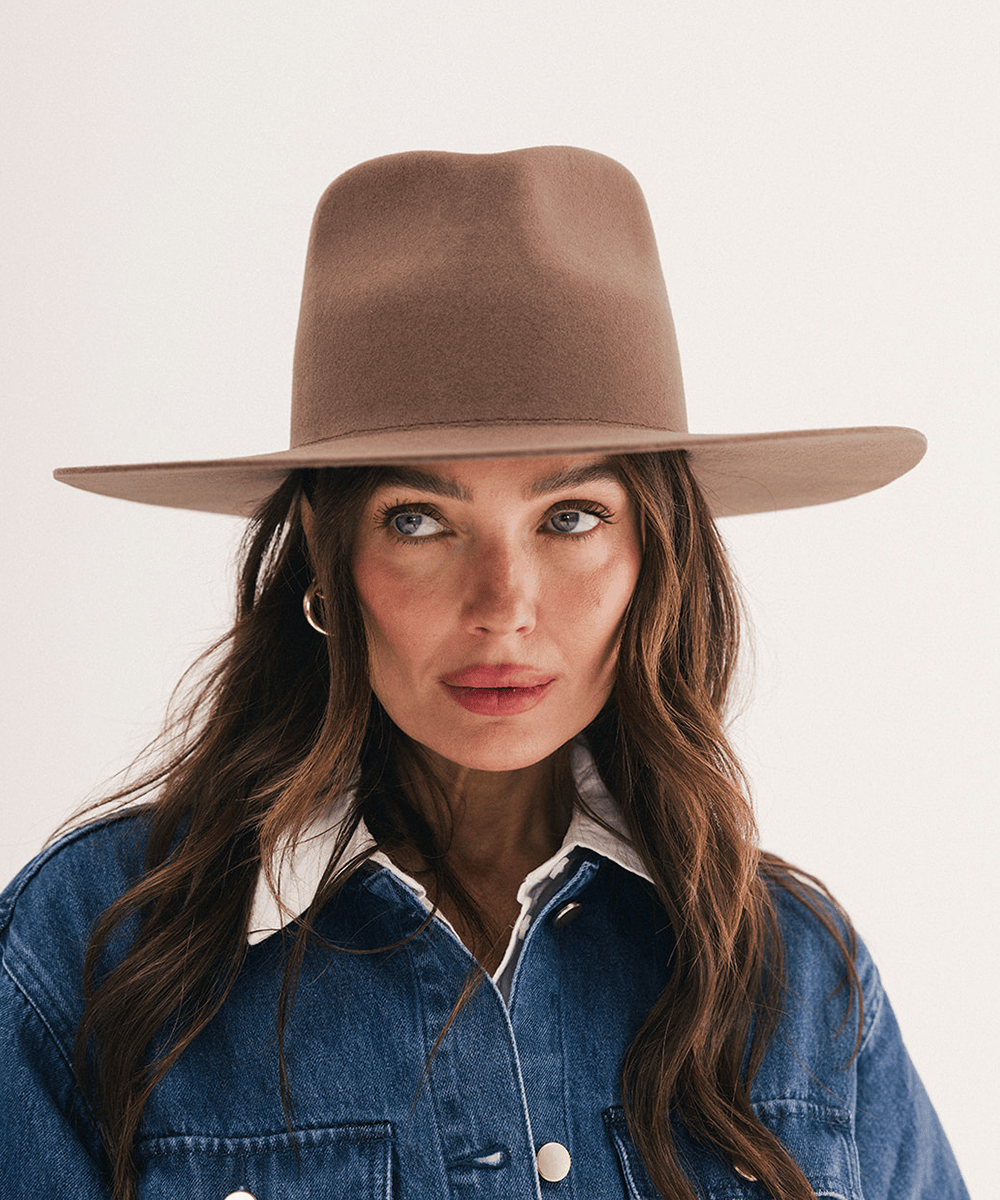 A woman wearing a wide-brimmed brown felt hat and a blue denim shirt, photographed against a clean off-white background #color_chocolate