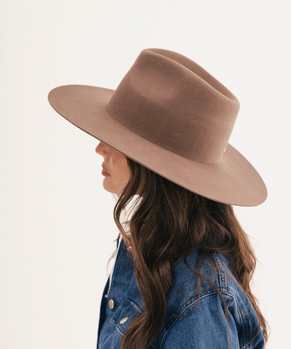 A woman in profile wearing a wide-brimmed felt hat in a warm taupe-brown color, paired with a blue denim jacket, against a clean white background #color_chocolate