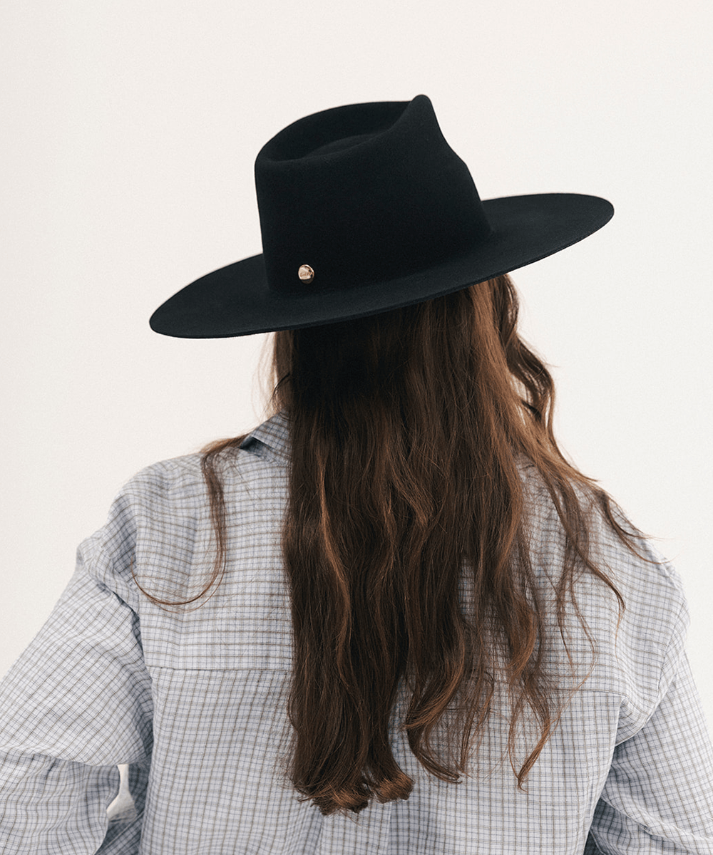 A person viewed from behind wearing a wide-brim black felt hat with a small gold button detail, a light blue and white checkered shirt, and long wavy brown hair against a plain off-white background #color_black