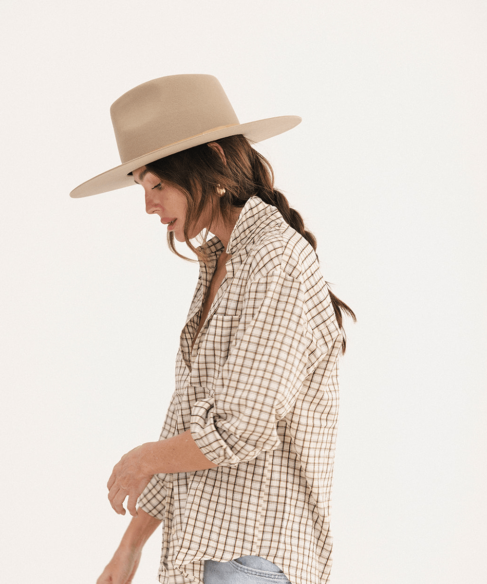 A woman wearing a beige wide-brim fedora hat and a beige and brown checkered button-down shirt, shown in profile against a plain white studio background #color_tan