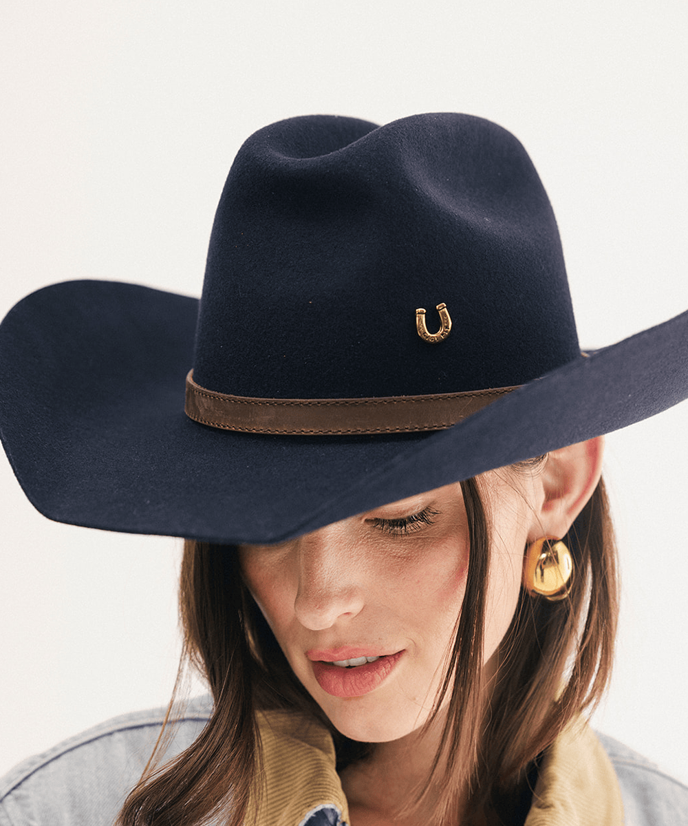 A close-up of a woman wearing a dark navy blue felt cowboy hat with a brown band and a small gold horseshoe emblem on the side, against a plain light background #color_navy