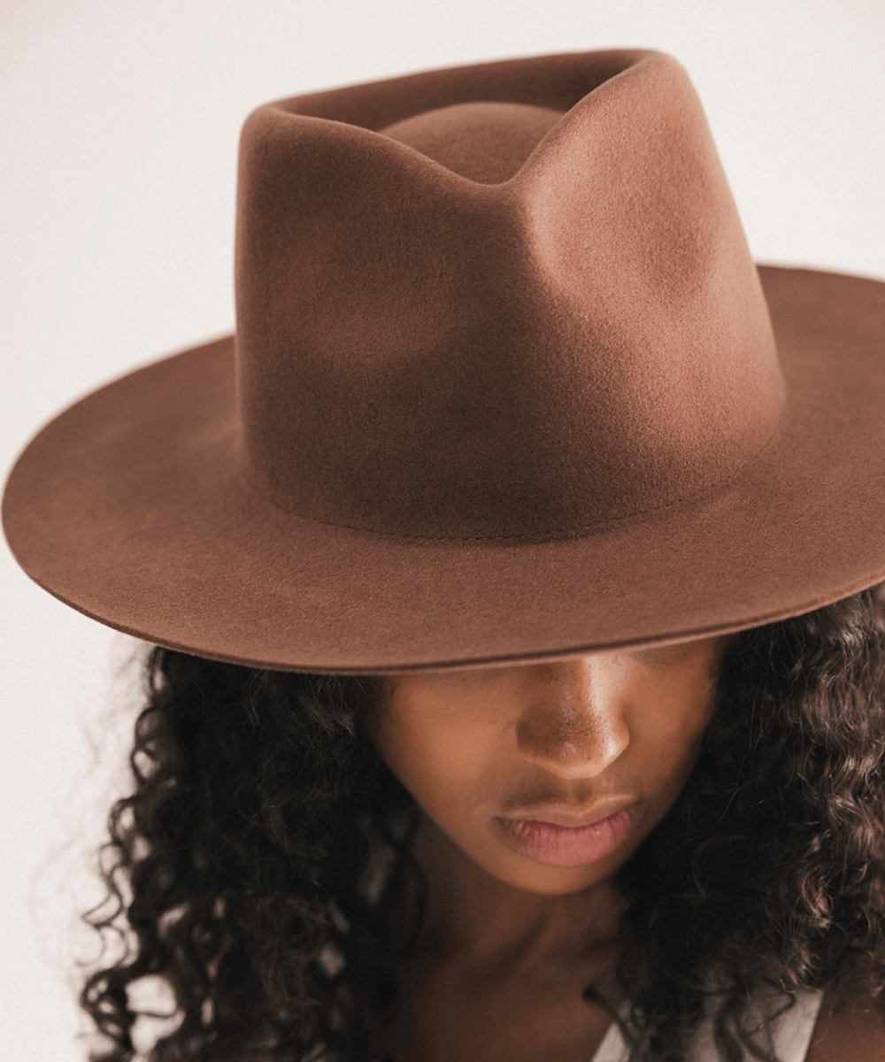 A close-up of a model wearing a brown felt wide-brim hat, showing the texture of the hat and her dark curly hair against a plain light background #color_dark oak