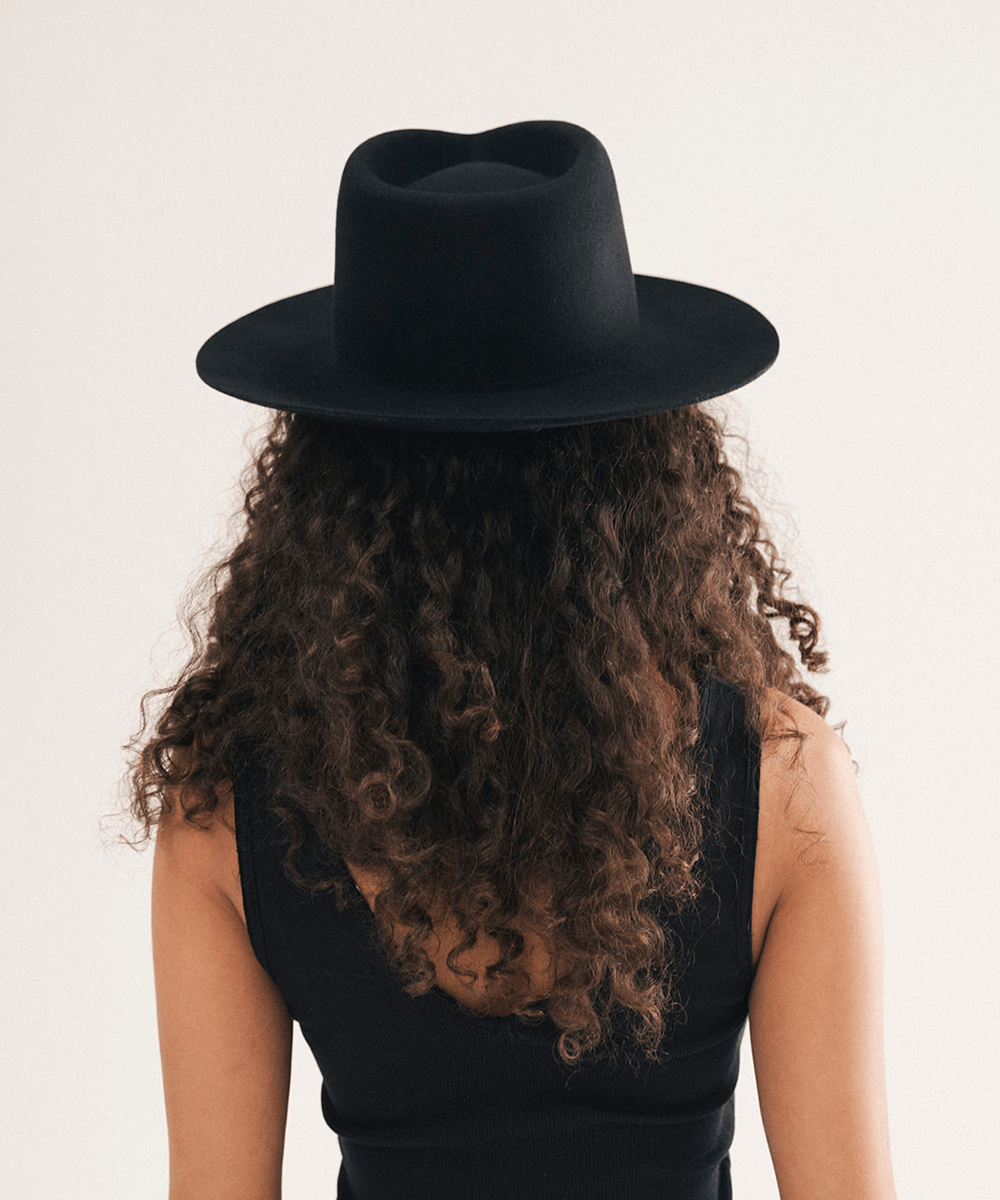 A person shown from the back wearing a black wide-brim fedora hat and a black sleeveless top, with long curly brown hair, against a plain light beige studio background #color_black