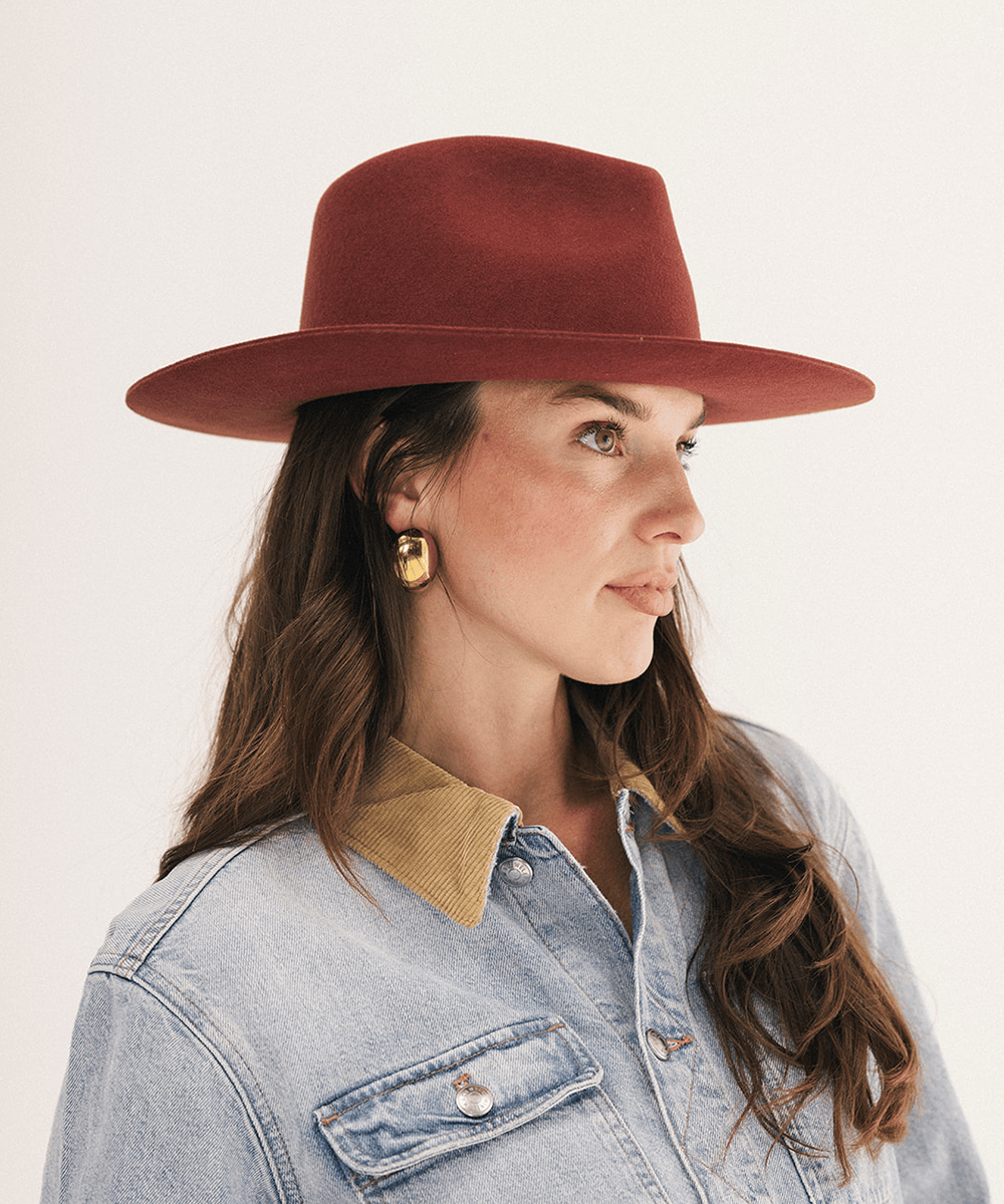 A woman in profile wearing a wide-brimmed rust-red felt hat, a light blue denim shirt with a tan corduroy collar, and gold geometric earrings, set against a plain off-white background #color_rusty red