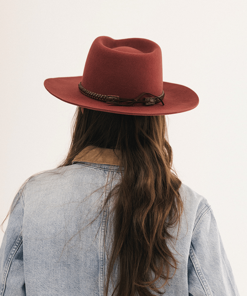 A person viewed from behind wearing a wide-brimmed rust-red felt hat with a dark braided band, a light blue denim jacket, and long brown hair against a plain off-white background #color_rusty red