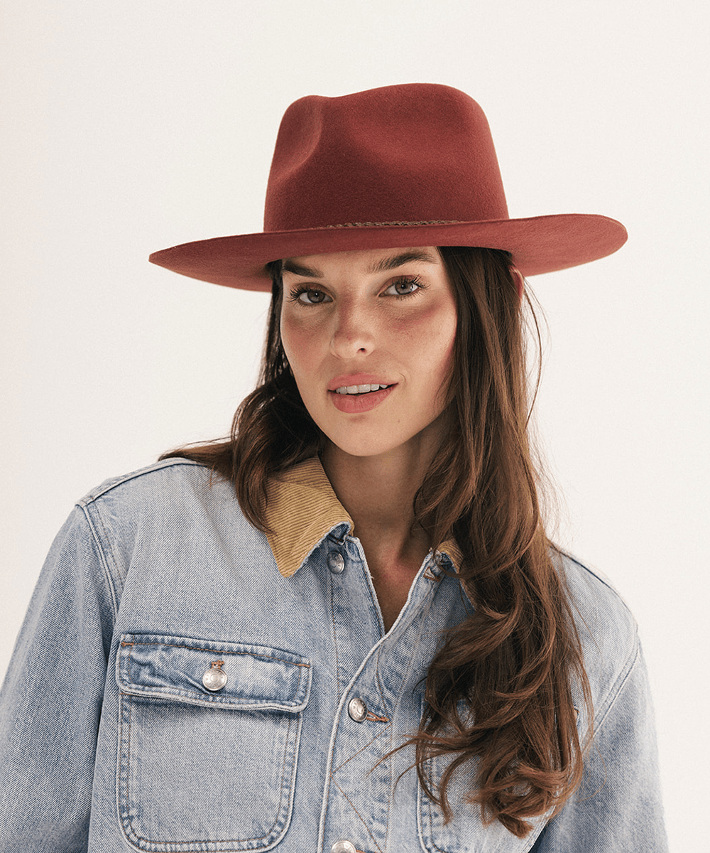 A woman wearing a rust-brown felt wide-brim hat and a light-wash denim shirt with a tan collar, photographed against a clean off-white studio background #color_rusty red