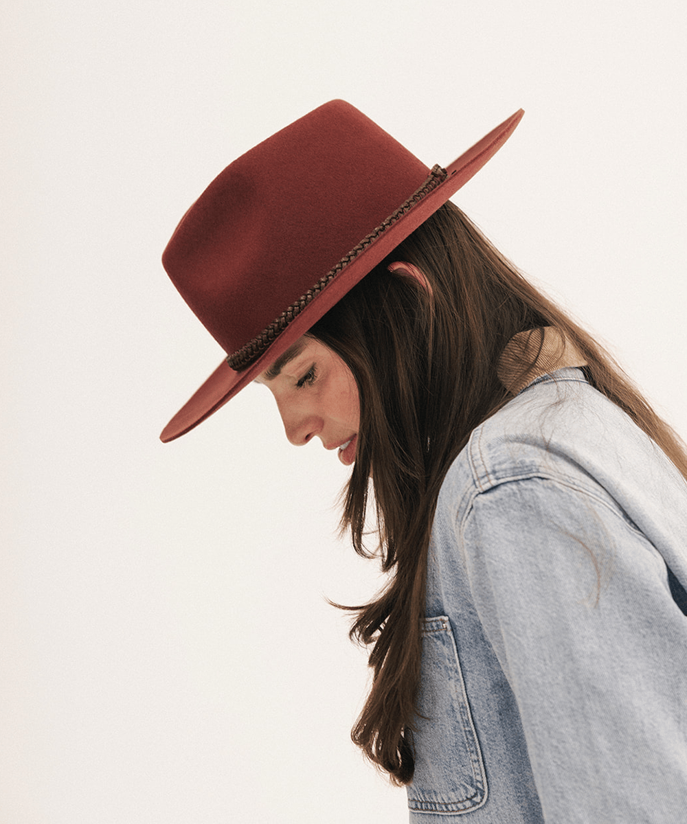 A woman in profile wearing a rust-red felt wide-brim hat with a braided band and a light blue denim jacket, set against a plain off-white background #color_rusty red