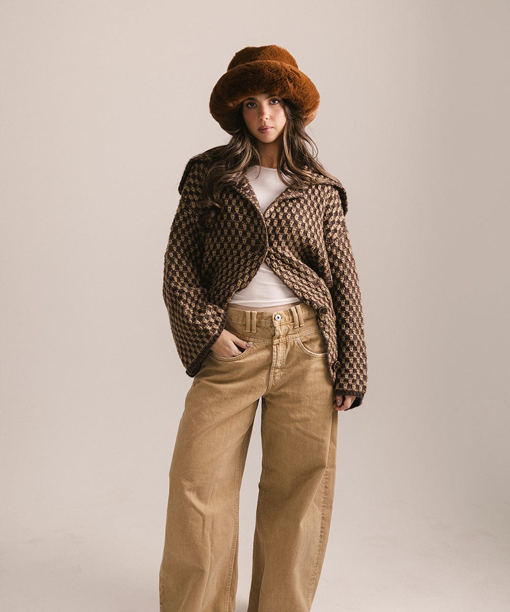 Woman wearing a patterned coat, white shirt, and beige pants with a brown hat on a plain background #color_brown