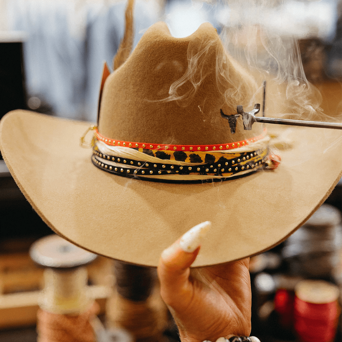 Person holding a brown cowboy hat with decorative band, blurred background at a custom hat bar event.