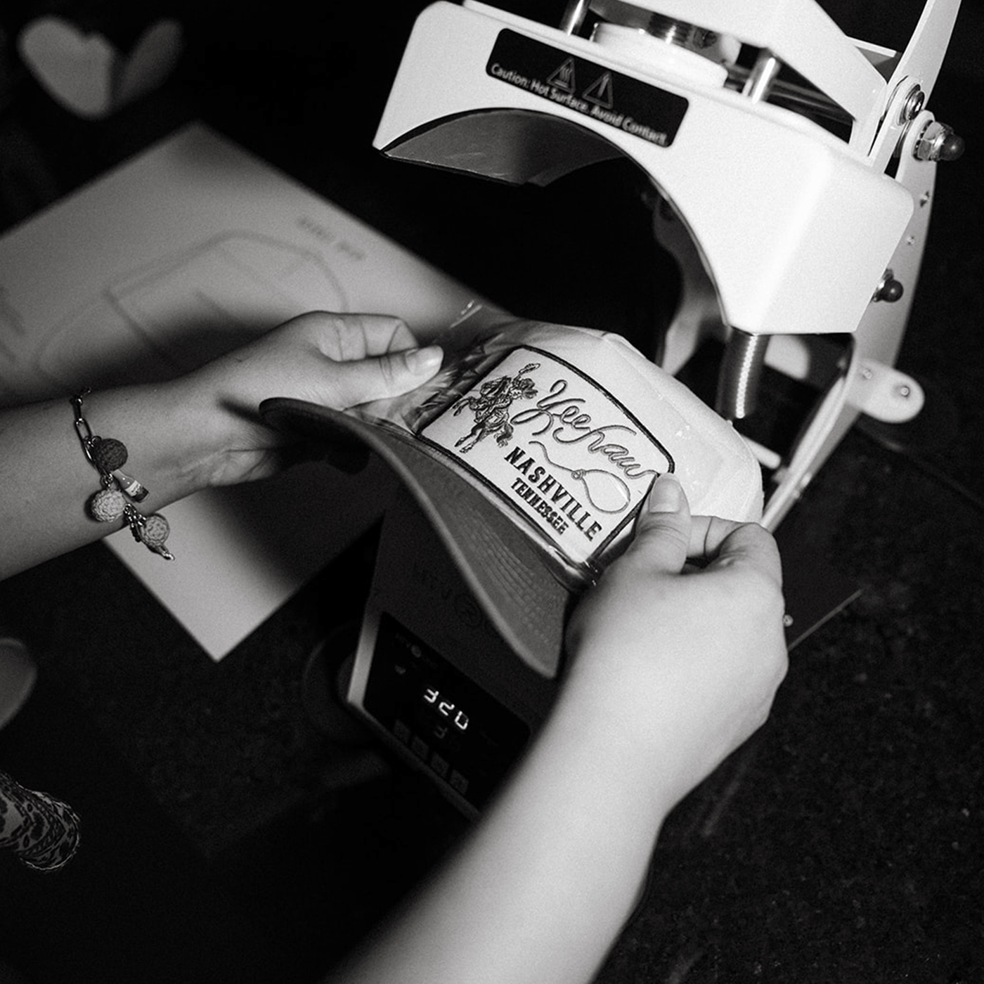 Person applying a design on the hat with a machine in a black and white setting at a custom hat bar event.