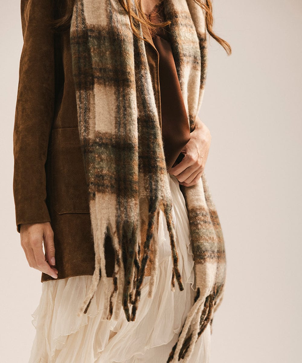 Person wearing a plaid scarf with fringe, brown coat, and white dress on a neutral background #color_cream - tan