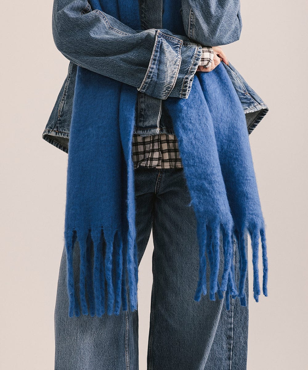 Person wearing a blue scarf with fringes, denim jacket, and jeans on a beige background #color_alpine blue