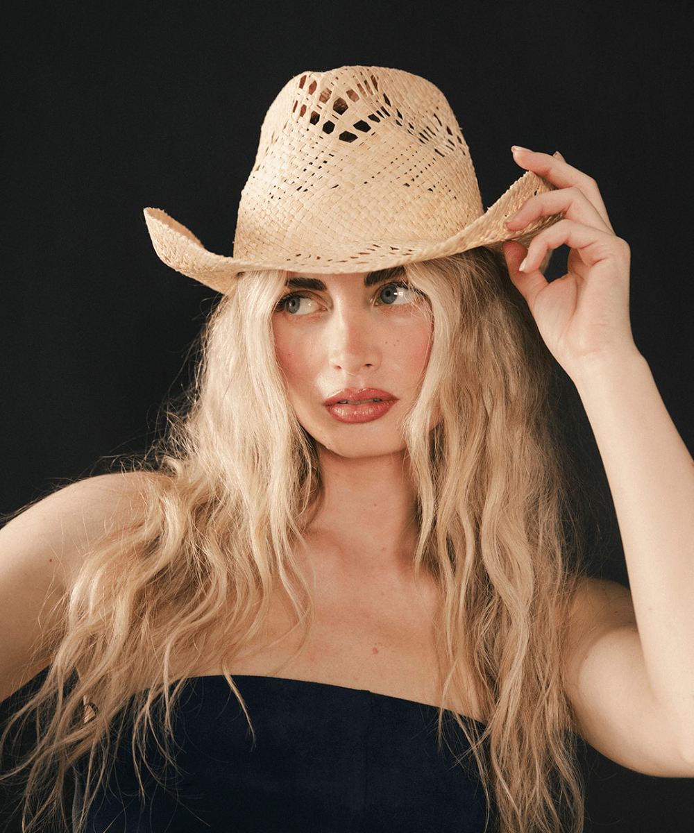 Woman wearing a natural colored straw cowboy hat and dark clothing against a dark background #color_natural
