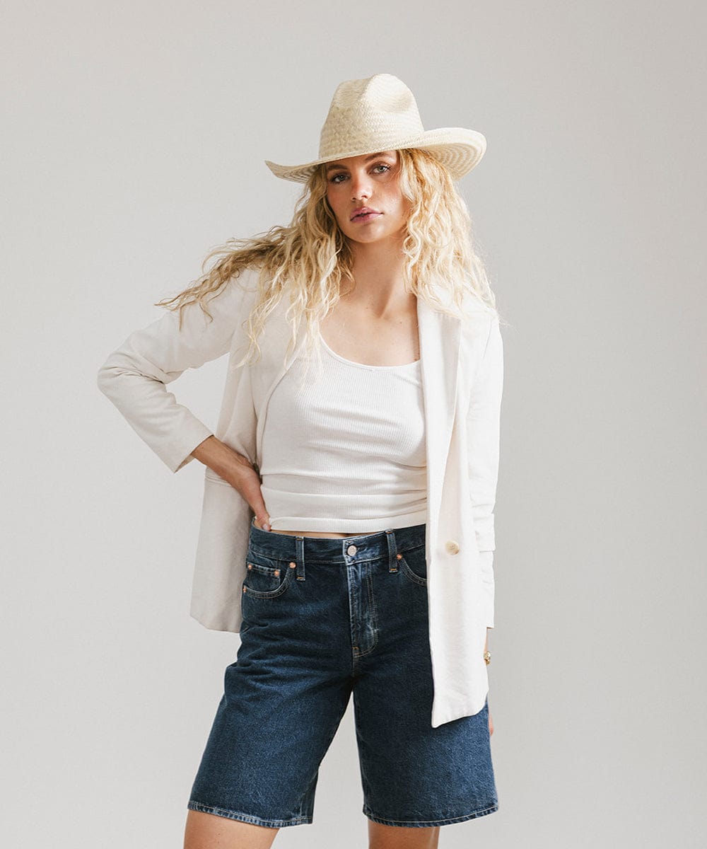 Woman wearing a natural colored straw cowboy hat, a white top with a white coat and denim shorts against a plain background  #color_natural