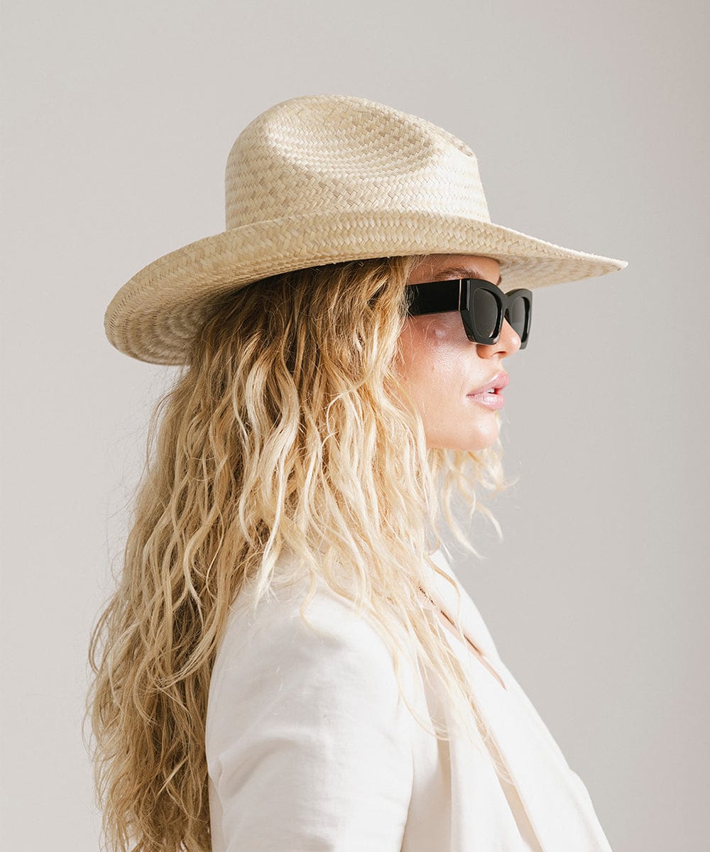 Woman wearing a natural colored straw cowboy hat, sunglasses and a white coat, facing sideways against a plain background  #color_natural