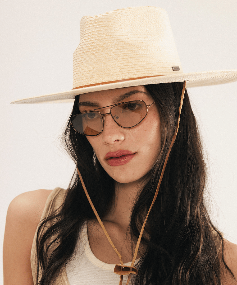Woman wearing an ivory colored palm straw sun hat with a teardrop fedora crown and a wide flat brim, featuring a brown genuine leather chinstrap on a white background #color_ivory