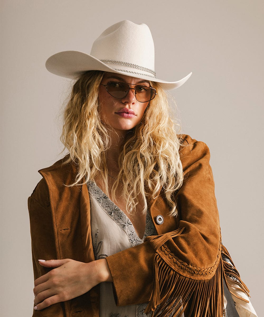 Woman wearing a cowboy hat, brown sunglasses and brown jacket with fringes against a neutral background #color_brown