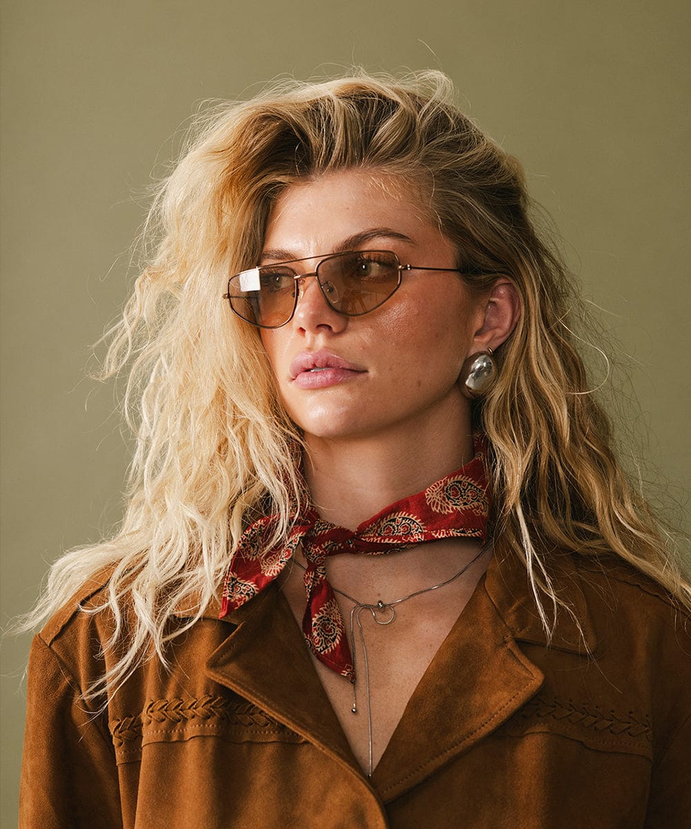 Woman wearing brown sunglasses and a brown jacket with a red scarf against a green background #color_brown