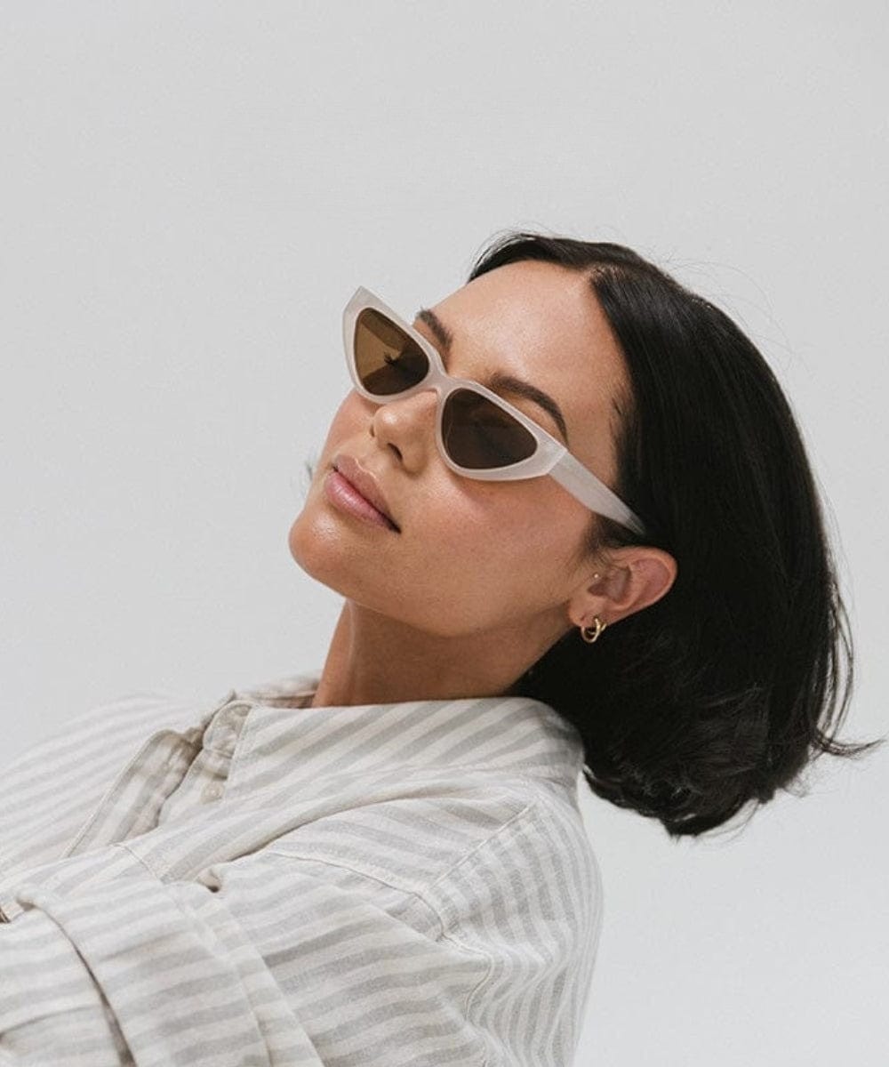 Woman wearing a light grey cat eye sunglasses and a white shirt with grey lines against a plain background #color_light grey