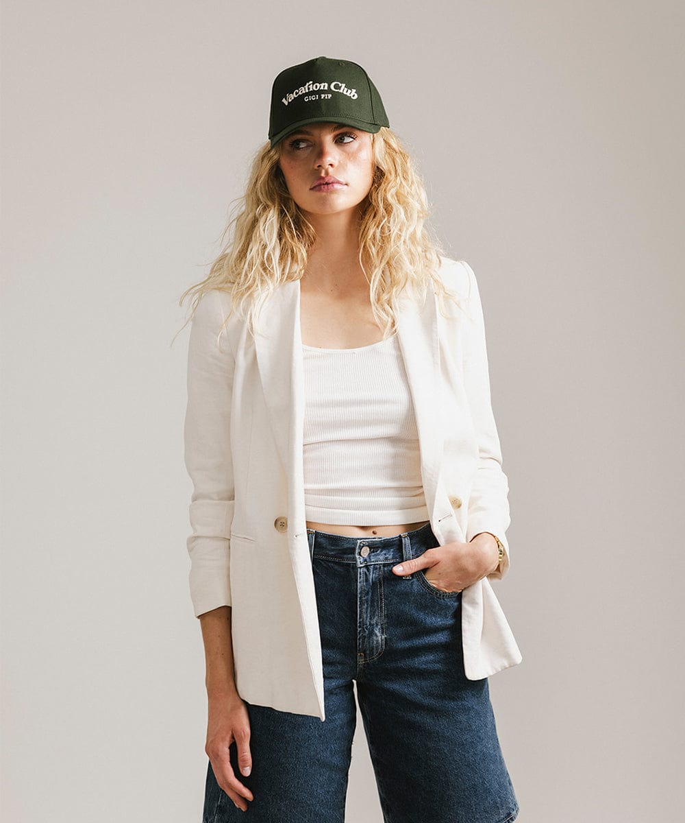 Woman wearing a dark green cap with 'Vacation Club' text, a white top with a white coat and denim shorts  against a plain background  #color_dark green
