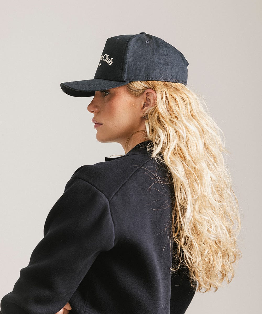 Woman wearing a navy cap with 'Vacation Club' text and a black shirt, facing sideways against a plain background  #color_navy