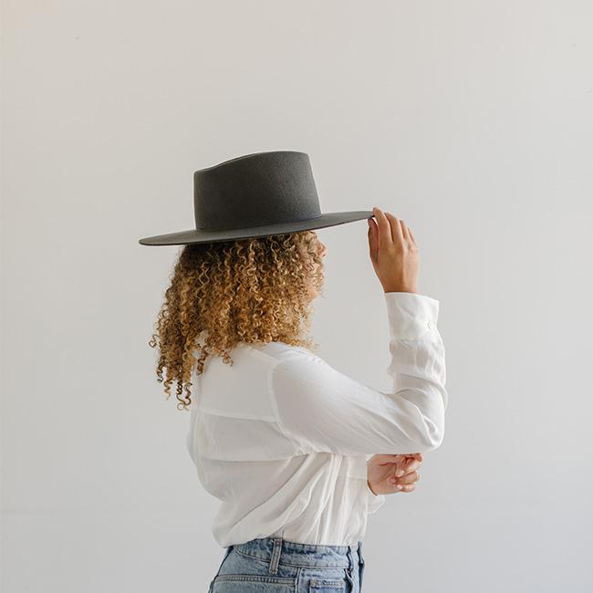 Woman wearing a dark grey fedora hat with a triangle crown, a white shirt and blue jeans, facing sideways against a plain background #color_dark grey