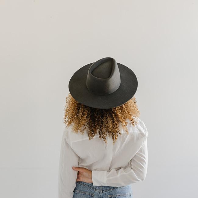Woman wearing a dark grey fedora hat with a triangle crown, a white shirt and blue jeans, facing behind against a plain background #color_dark grey