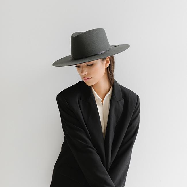 Woman wearing a dark grey fedora hat with a triangle crown and hat band and a white shirt with a black blazer, posing against a plain background #color_dark grey