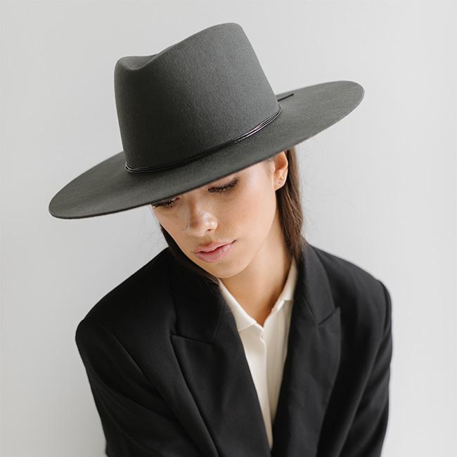 Woman wearing a dark grey fedora hat with a triangle crown and hat band and a white shirt with a black blazer against a plain background #color_dark grey