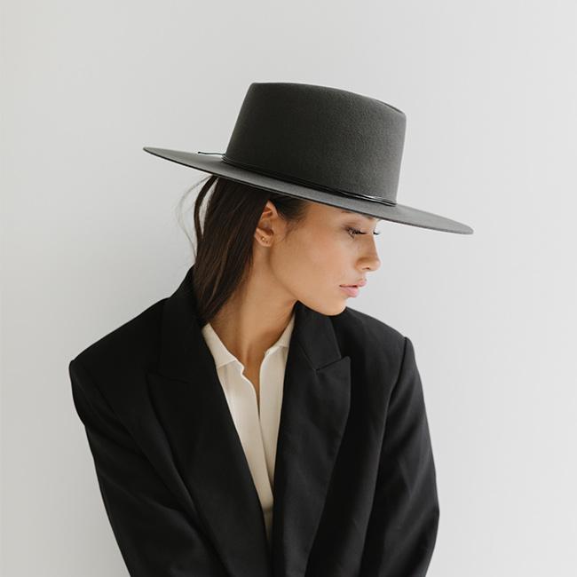 Woman wearing a dark grey fedora hat with a triangle crown and hat band and a white shirt with a black blazer, looking sideways against a plain background #color_dark grey