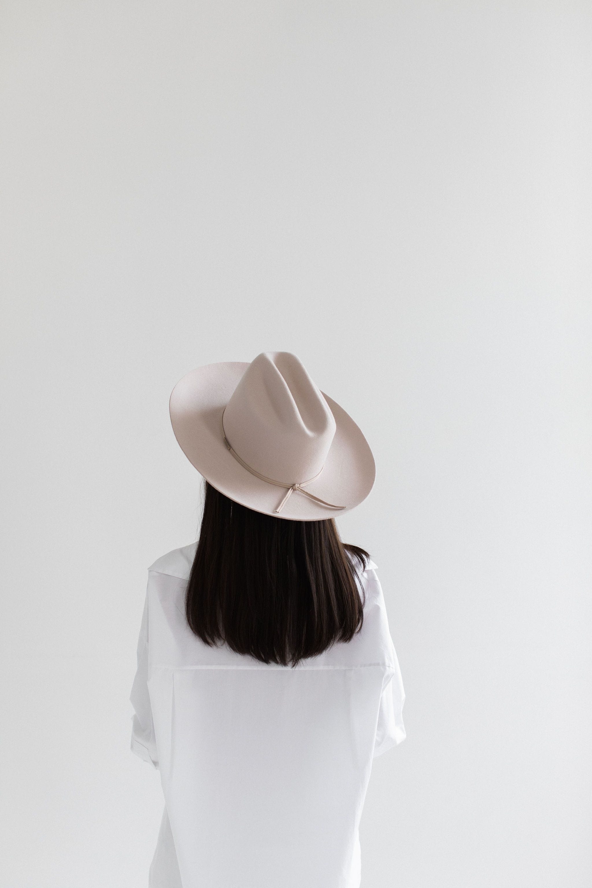Woman wearing a ivory colored cowboy hat with a hat band and a white shirt, facing behind against a plain background #color_ivory