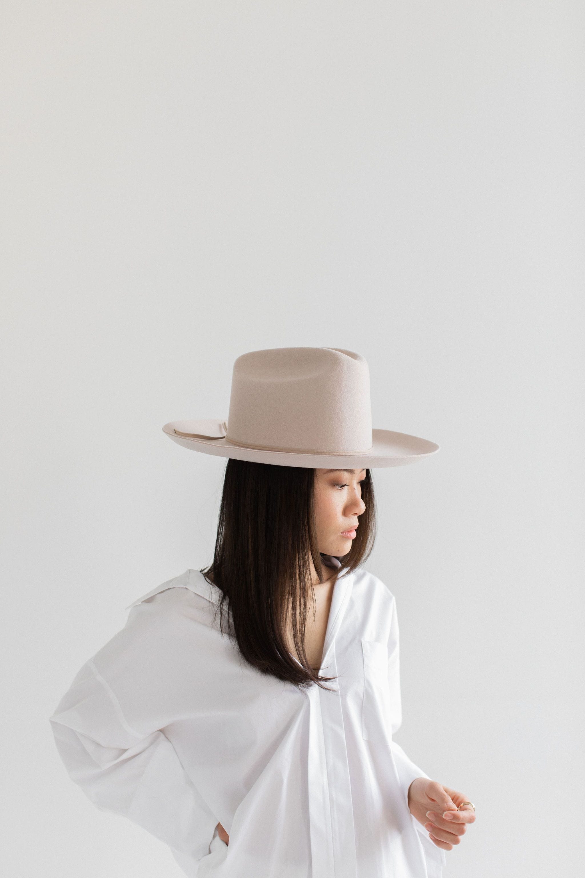 Woman wearing a ivory colored cowboy hat with a hat band and a white shirt, posing sideways against a plain background #color_ivory
