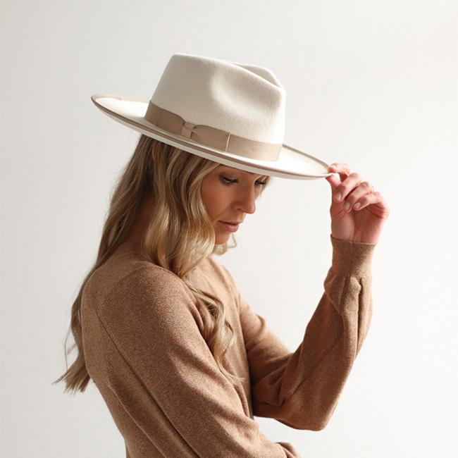 Woman wearing an off white and taupe colored fedora hat with a taupe band and a brown sweatshirt, looking sideways against a plain background #color_white-taupe