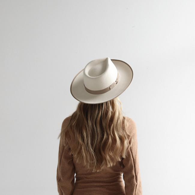 Woman wearing an off white and taupe colored fedora hat with a taupe band and a brown sweatshirt, facing behind against a plain background #color_white-taupe