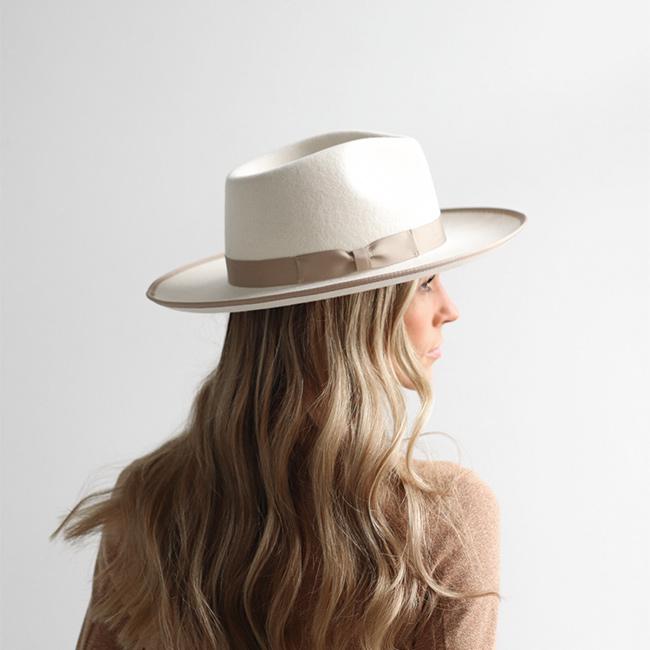 Woman wearing an off white and taupe colored fedora hat with a taupe band and a brown sweatshirt, facing sideways against a plain background #color_white-taupe