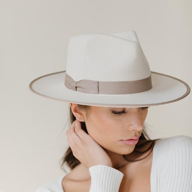 Woman wearing an off white and taupe colored fedora hat with a taupe band and a white top against a plain background #color_white-taupe