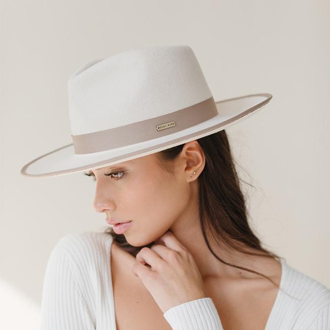 Woman wearing an off white and taupe colored fedora hat with a taupe band and a white top, looking sideways against a plain background #color_white-taupe