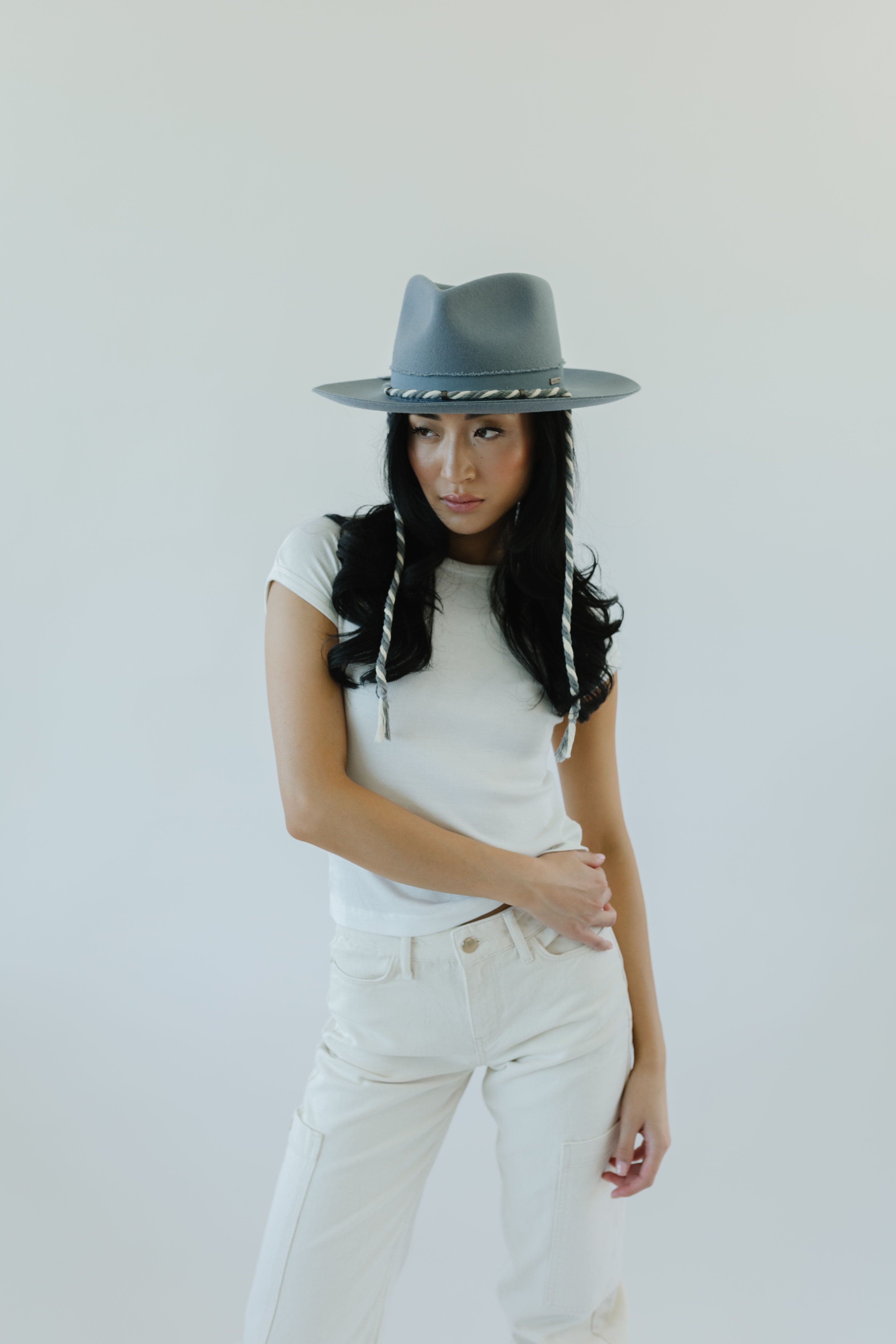 Woman wearing a denim colored wide brim hat with a braided chin strap, a white t-shirt and white pants against a plain background  #color_denim