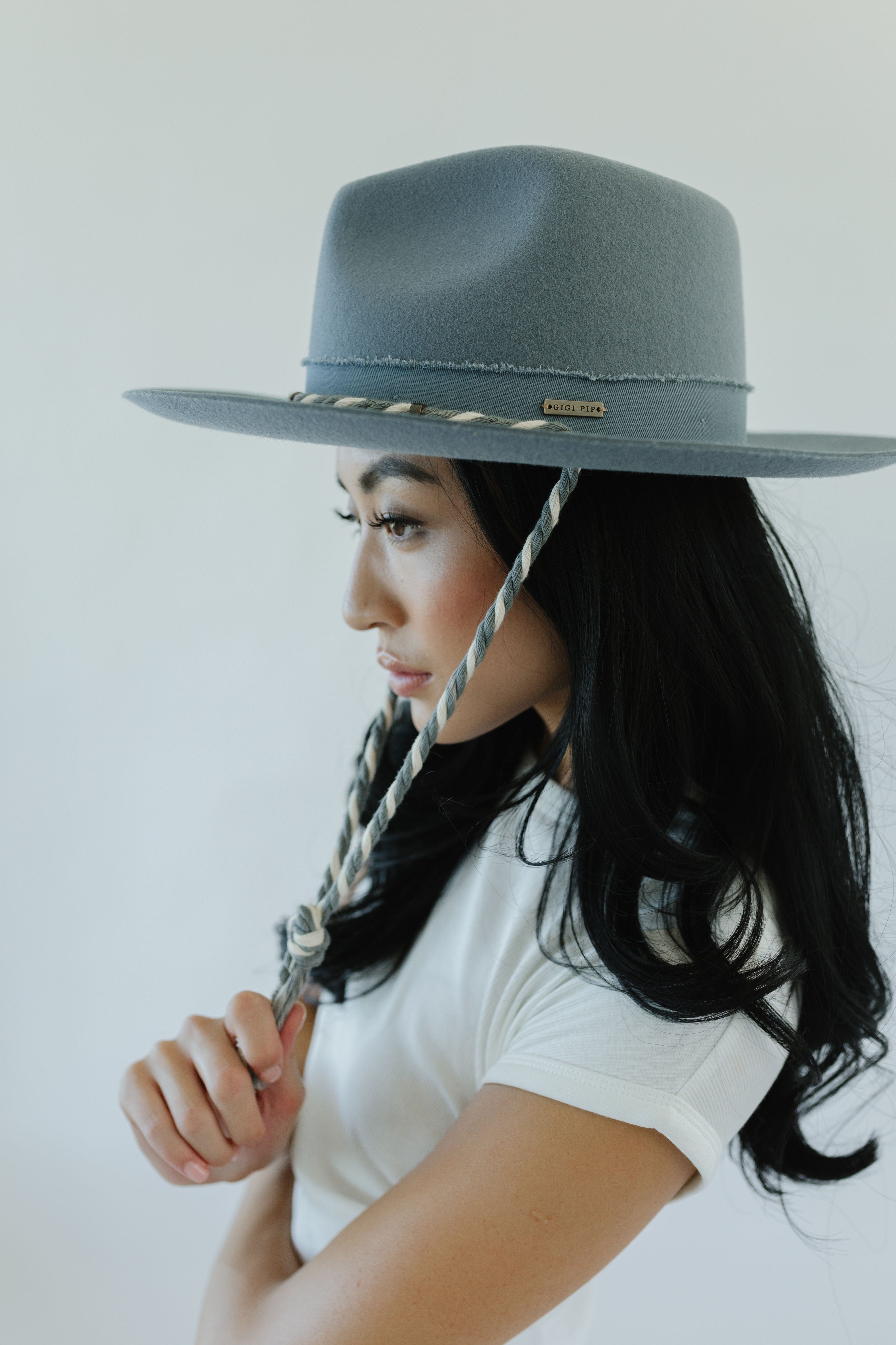 Woman wearing a denim colored wide brim hat with a braided chin strap and  a white t-shirt, facing sideways against a plain background  #color_denim