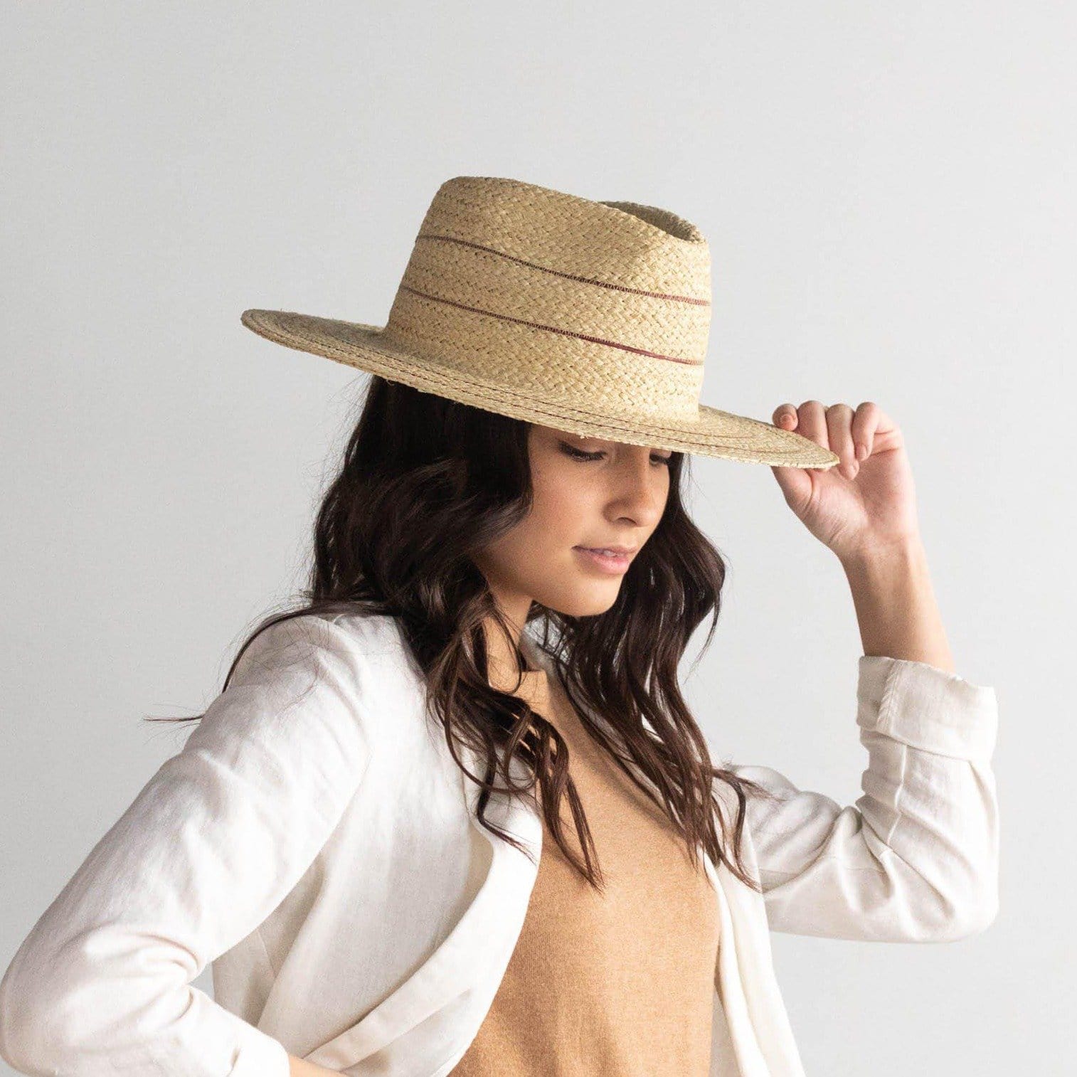 Woman wearing a natural colored straw fedora hat with stripes and a brown t-shirt with a cream jacket, looking sideways against a plain background #color_natural with stripes