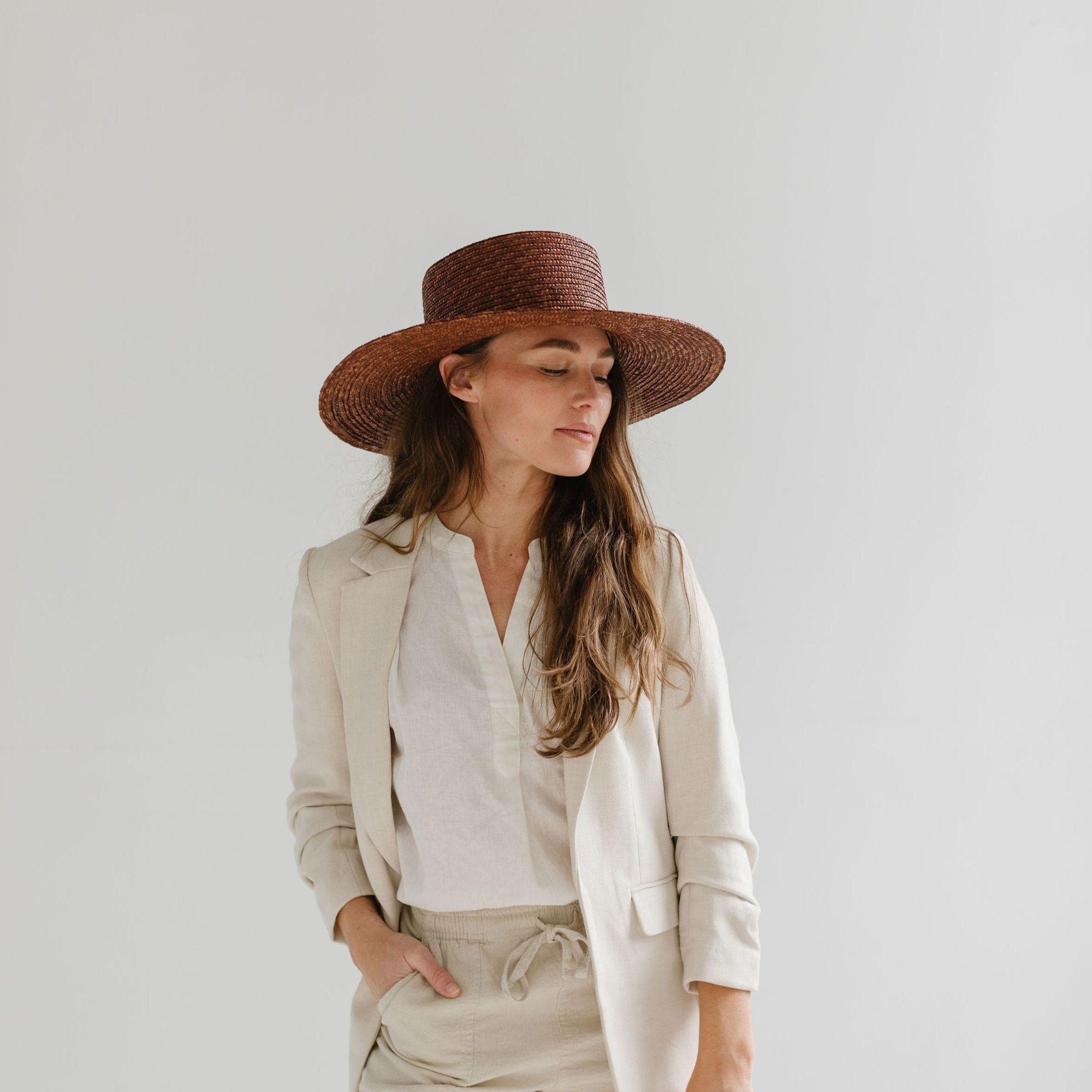 Woman wearing a brown boater-style straw hat with a flat brim and a hat band, a white shirt with a beige blazer and beige shorts , posing against a plain background #color_brown