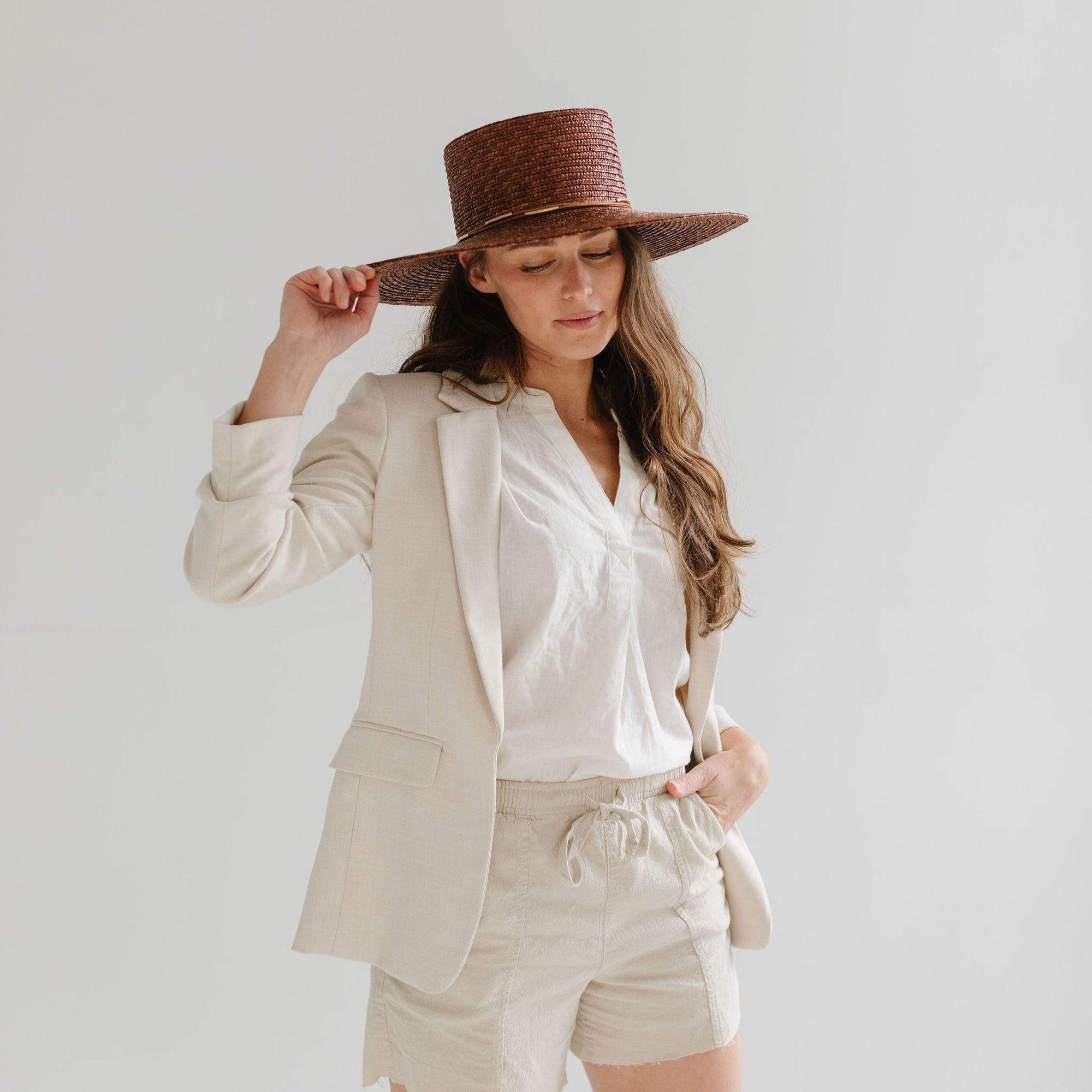 Woman wearing a brown boater-style straw hat with a flat brim and a hat band, a white shirt with a beige blazer and beige shorts against a plain background #color_brown