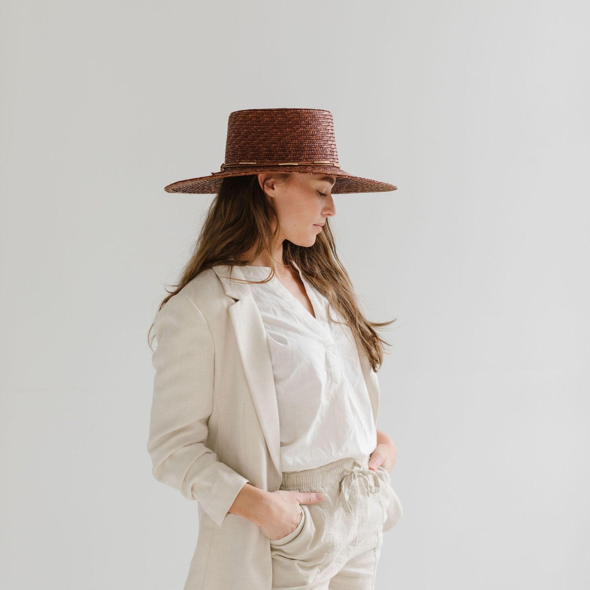 Woman wearing a brown boater-style straw hat with a flat brim and a hat band, a white shirt with a beige blazer and beige shorts, looking sideways against a plain background #color_brown