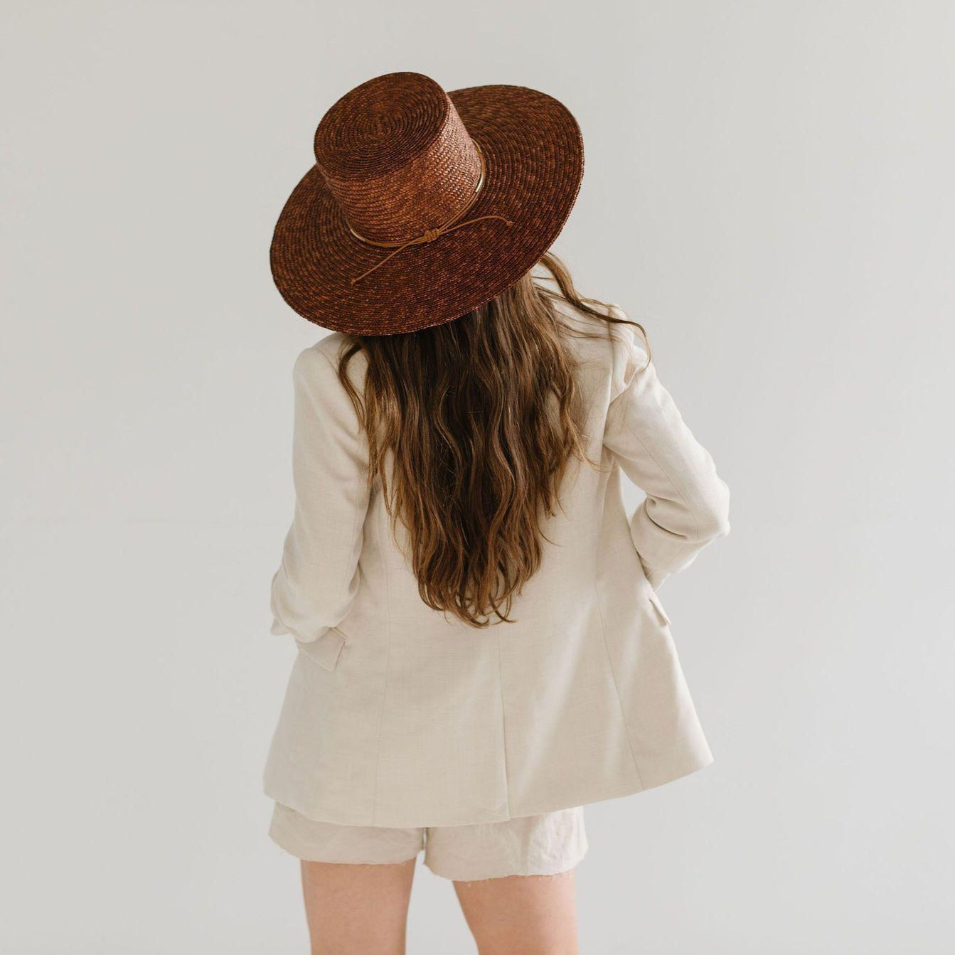 Woman wearing a brown boater-style straw hat with a flat brim and a hat band, a white shirt with a beige blazer and beige shorts, facing behind against a plain background #color_brown